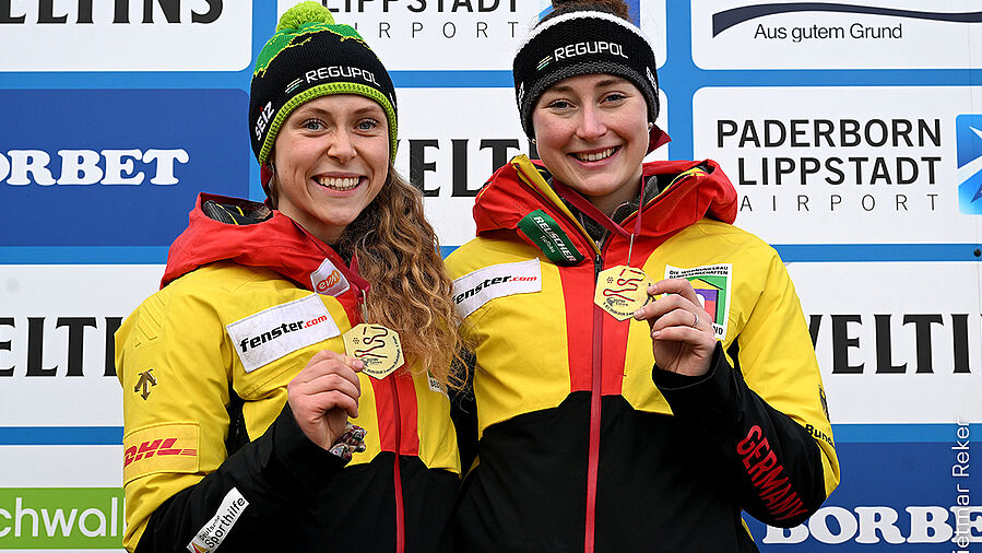 Charlotte Candrix und Lena Brunnhübner mit Goldmedaillen auf dem Podest bei einer Siegerehrung Charlotte Candrix mit Lena Brunnhübner als Siegerinnen im Zweierbob mit Goldmedaillen und REGUPOL Mützen
